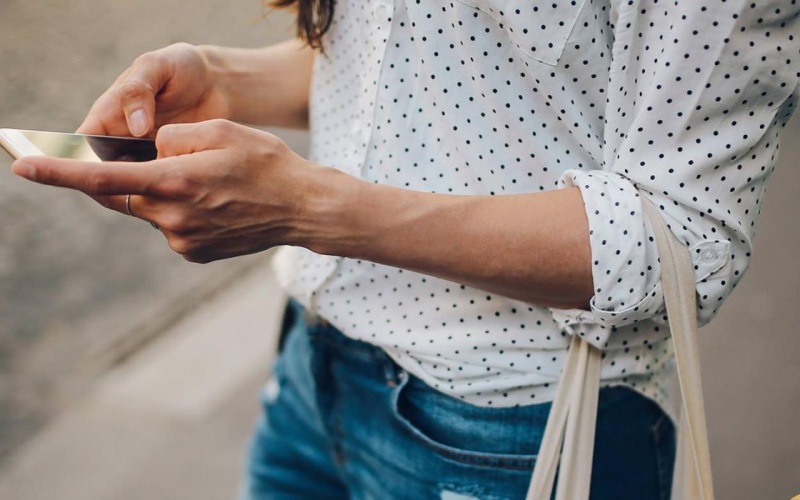 woman walks down the street and texts with a bag of groceries on her arm